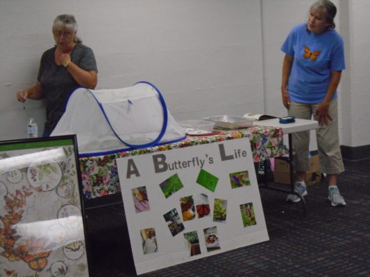 Two women standing beside a table displaying Monarch caterpillars, butterflies and milk weed. Also is a poster showing the life cycle of the Monarch Butterfly