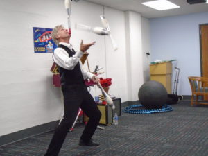 man in vest balancing a bowling pin on his chin, while juggling 3 other pins
