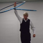 man in vest twirling 2 hula hoops above his head on his right arm